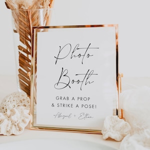 May include: A gold-framed sign with the words "Photo Booth" in elegant script. Below, it reads "Grab a prop & strike a pose!" with names Abigail + Ethan. The sign is on a white surface.