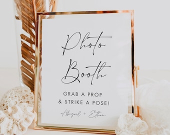 May include: A gold-framed sign with the words "Photo Booth" in elegant script. Below, it reads "Grab a prop & strike a pose!" with names Abigail + Ethan. The sign is on a white surface.