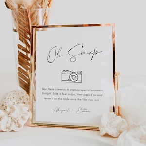 May include: A gold framed sign with the words "Oh Snap" in elegant script. Below is a camera illustration and text encouraging guests to take photos. The sign is a wedding or event decoration, with the names "Abigail + Ethan" at the bottom.