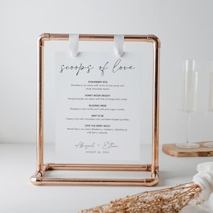 Könnte beinhalten: Ein kupferfarbener Metallrahmen hält ein weißes Schild mit der Aufschrift "scoops of love". Das Schild listet Eissorten wie Strawberry Kiss und Honey Moon Delight auf. Weiße Bänder sichern das Schild.