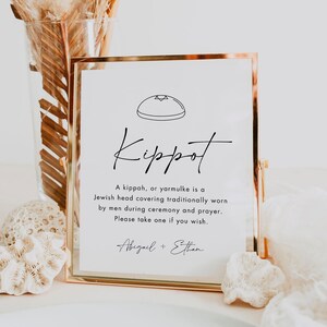 May include: A gold-framed sign with the word "Kippot" in elegant script. The sign includes a description of a kippah, or yarmulke, and the text "Please take one if you wish." The names "Abigail + Ethan" are at the bottom.