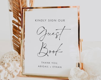 May include: A gold-framed sign with the words "KINDLY SIGN OUR Guest Book" in black script. Below, it reads "THANK YOU, ABIGAIL + ETHAN." The sign is on a white surface, with decorative elements in the background.