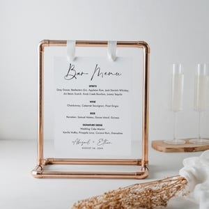 May include: A copper-colored metal bar menu sign with a white paper insert. The sign reads "Bar Menu" in elegant script, listing spirits, wine, beer, and a signature drink. The sign is held in place with white ribbons. The names "Abigail + Ethan" and the date "August 24, 20XX" are also displayed.