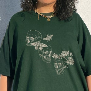 May include: A dark green t-shirt with a graphic print of three skulls with flowers and a butterfly. The skulls are white and the flowers are black.
