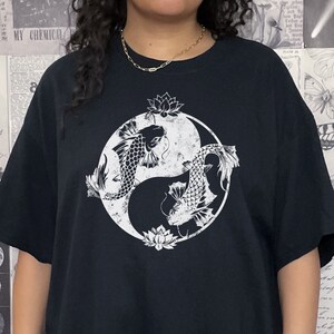 May include: A black t-shirt with a white yin yang symbol featuring two fish and floral designs. The model is wearing the shirt two sizes up from their normal size.