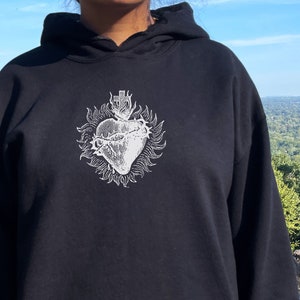 May include: Black hooded sweatshirt with a white graphic of a heart with thorns and a crown of thorns. The model is wearing a size up from her normal size.