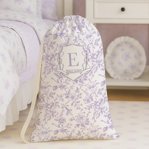 May include: A white drawstring laundry bag with a floral pattern in lavender. The bag features a shield with the letter "E" and the name "EMILY MARIE". The bag is standing in front of a bed with matching bedding.