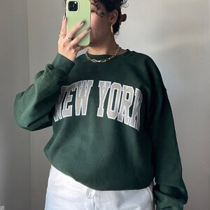 May include: A dark green sweatshirt with the words "NEW YORK" in white and beige lettering. The model is wearing the sweatshirt with white shorts.