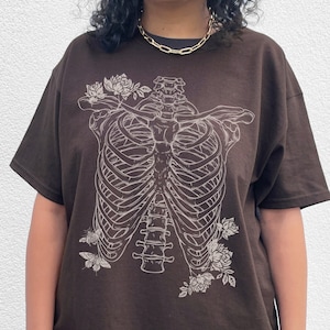 May include: A brown t-shirt with a white graphic of a ribcage with flowers. The model is wearing the shirt two sizes up from her normal size. "THE MODEL IS WEARING 2 SIZES UP FROM HER NORMAL SIZE"