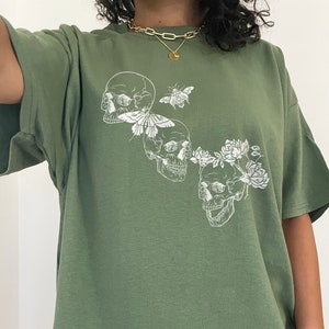 Puede incluir: Una camiseta verde de gran tama&ntilde;o con un estampado gr&aacute;fico blanco de tres calaveras con flores y una mariposa. La modelo lleva la camiseta dos tallas m&aacute;s grande que su talla normal.