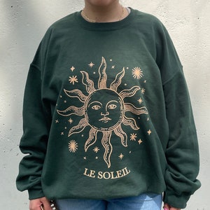 May include: A dark green sweatshirt with a gold sun graphic and the text "LE SOLEIL" printed on the front.