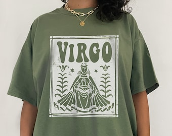 Puede incluir: Camiseta verde de gran tamaño con un gráfico blanco de un signo del zodíaco Virgo. El gráfico presenta a una mujer con una corona sentada en un trono con flores y estrellas.