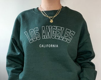 Puede incluir: Una sudadera verde oscuro con el texto "Los Ángeles, California" en letras blancas delineadas.