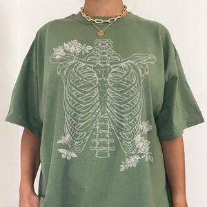 K&ouml;nnte beinhalten: Ein gr&uuml;nes Oversize-T-Shirt mit einem wei&szlig;en Aufdruck eines Brustkorbs und Blumen. Das Model tr&auml;gt das Shirt zwei Gr&ouml;&szlig;en gr&ouml;&szlig;er als seine normale Gr&ouml;&szlig;e.
