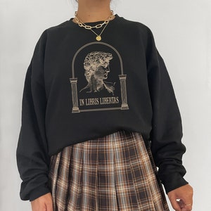 May include: Black sweatshirt with a graphic of a man's face in an archway with the text "In Libris Libertas". The model is wearing a brown and white plaid skirt.