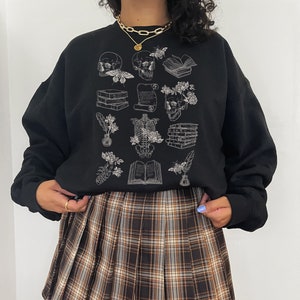 May include: Black sweatshirt with a white graphic print of skulls, books, flowers, and other gothic-inspired imagery. The sweatshirt is paired with a brown and white plaid pleated skirt.
