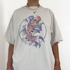 May include: A light gray oversized t-shirt with a graphic print of a red and blue koi fish swimming in a circle of water. The model is wearing the shirt two sizes up from her normal size. "THE MODEL IS WEARING 2 SIZES UP FROM HER NORMAL SIZE" is printed on the bottom of the image.