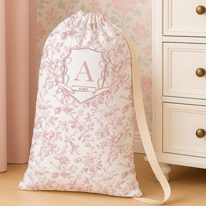 May include: A white drawstring laundry bag with a pink floral pattern and a large letter "A" inside a crest. The bag has a beige strap and is set against a floral wallpaper background.