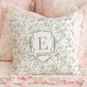 May include: A decorative pillow with a floral pattern and a gray monogrammed crest featuring the letter "E" and the name "Emily Marie". The pillow is surrounded by other pillows and bedding in shades of pink and white.