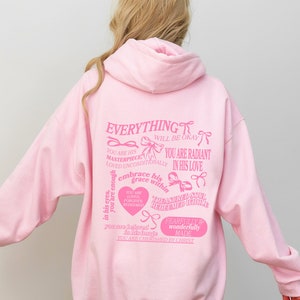 May include: Pink hoodie with a graphic print of inspirational text and bows. The text includes "Everything will be okay", "You are his masterpiece", "You are radiant in his love", "Embrace his grace within", "You are loved, forgiven, redeemed whole", "Fearfully and wonderfully made", and "You are cherished by Christ".