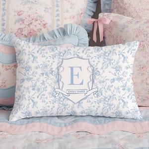 May include: A collection of decorative pillows in shades of blue, pink, and white. The pillows feature floral patterns and a monogrammed pillow with the letter "E" and the name "Emily Marie". The bedding has a romantic, vintage aesthetic.