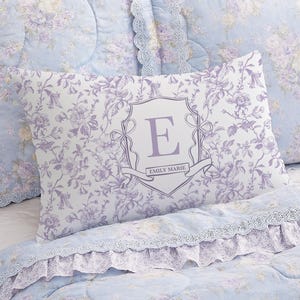May include: A light blue floral patterned pillow with a decorative monogram in a shield shape. The pillow has a white background with a purple floral design. The monogram is the letter "E" in purple, with the name "EMILY MARIE" below it.