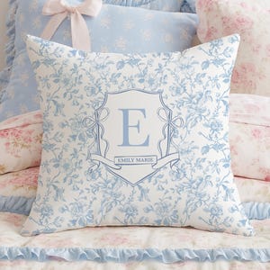 May include: A square cushion with a floral pattern in white and pale blue. The cushion features a central crest design with the letter "E" and the name "EMILY MARIE" in pale blue. Other cushions and bedding are visible in the background.