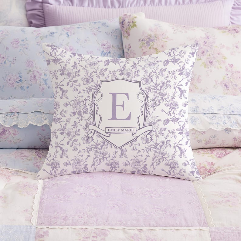 May include: A square decorative pillow with a floral pattern in shades of lavender and white. The pillow features a central crest design with the letter "E" and the name "EMILY MARIE" on a banner. The pillow is surrounded by other floral-patterned bedding.
