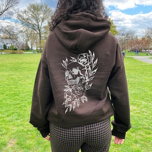 Puede incluir: Sudadera con capucha marr&oacute;n con un dise&ntilde;o de calavera blanca y flores en la espalda. La modelo lleva su talla normal.