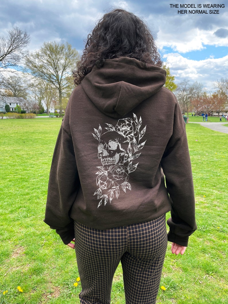 Puede incluir: Sudadera con capucha marr&oacute;n con un dise&ntilde;o de calavera blanca y flores en la espalda. La modelo lleva su talla normal.