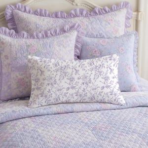 May include: A bed set featuring a quilted comforter and several pillows in shades of lavender and white. The pillows have ruffled edges and floral patterns. The comforter and pillows have a soft, romantic aesthetic.