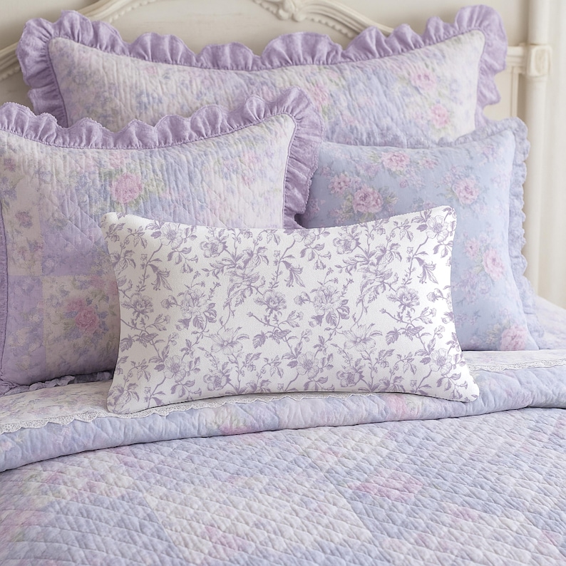 May include: A bed set featuring a quilted comforter and several pillows in shades of lavender and white. The pillows have ruffled edges and floral patterns. The comforter and pillows have a soft, romantic aesthetic.