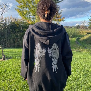 May include: Black hooded sweatshirt with a large rhinestone design of angel wings on the back. The model is wearing the sweatshirt two sizes up from her normal size.