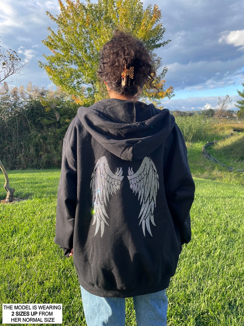 May include: Black hooded sweatshirt with a large rhinestone design of angel wings on the back. The model is wearing the sweatshirt two sizes up from her normal size.