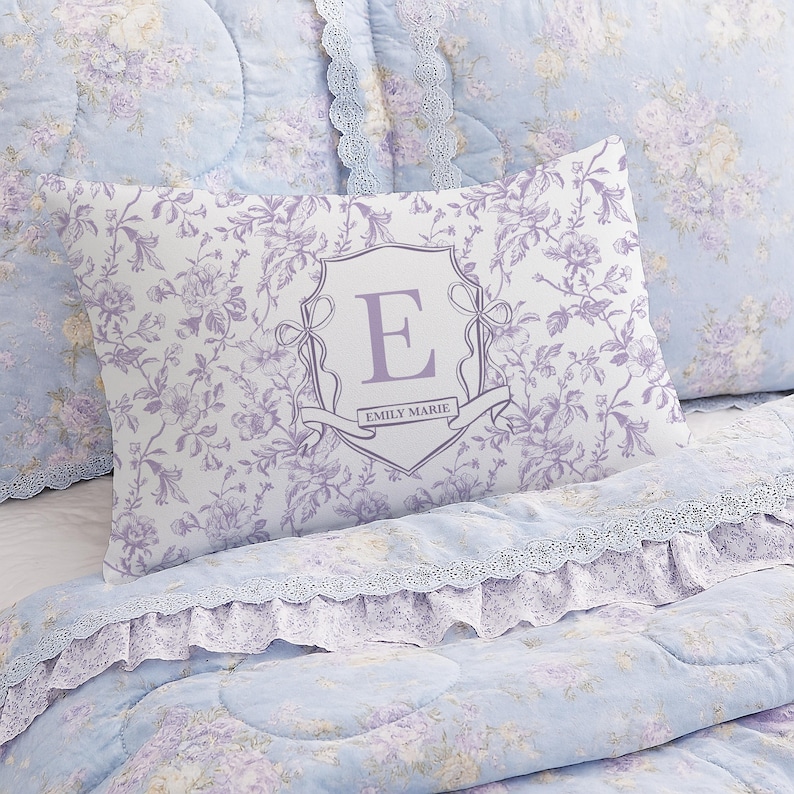 May include: A light blue floral patterned pillow with a decorative monogram in a shield shape. The pillow has a white background with a purple floral design. The monogram is the letter "E" in purple, with the name "EMILY MARIE" below it.