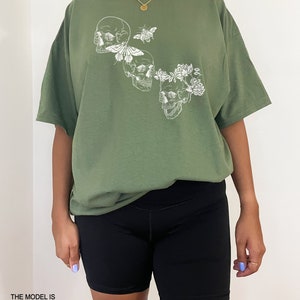 Puede incluir: Una camiseta verde de gran tama&ntilde;o con un estampado gr&aacute;fico blanco de tres calaveras con flores y una mariposa. La modelo lleva la camiseta dos tallas m&aacute;s grande que su talla habitual.