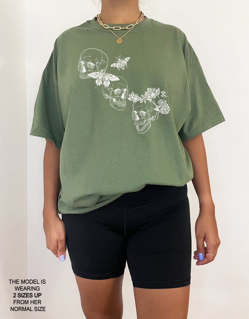 Puede incluir: Una camiseta verde de gran tama&ntilde;o con un estampado gr&aacute;fico blanco de tres calaveras con flores y una mariposa. La modelo lleva la camiseta dos tallas m&aacute;s grande que su talla habitual.