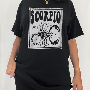 May include: Black oversized t-shirt with a white graphic of a scorpion and the word "SCORPIO" in a vintage style font.