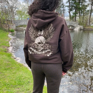 May include: Brown hooded sweatshirt with a skull graphic and the text "Lucky One" on the back. The model is wearing brown and black checkered pants.