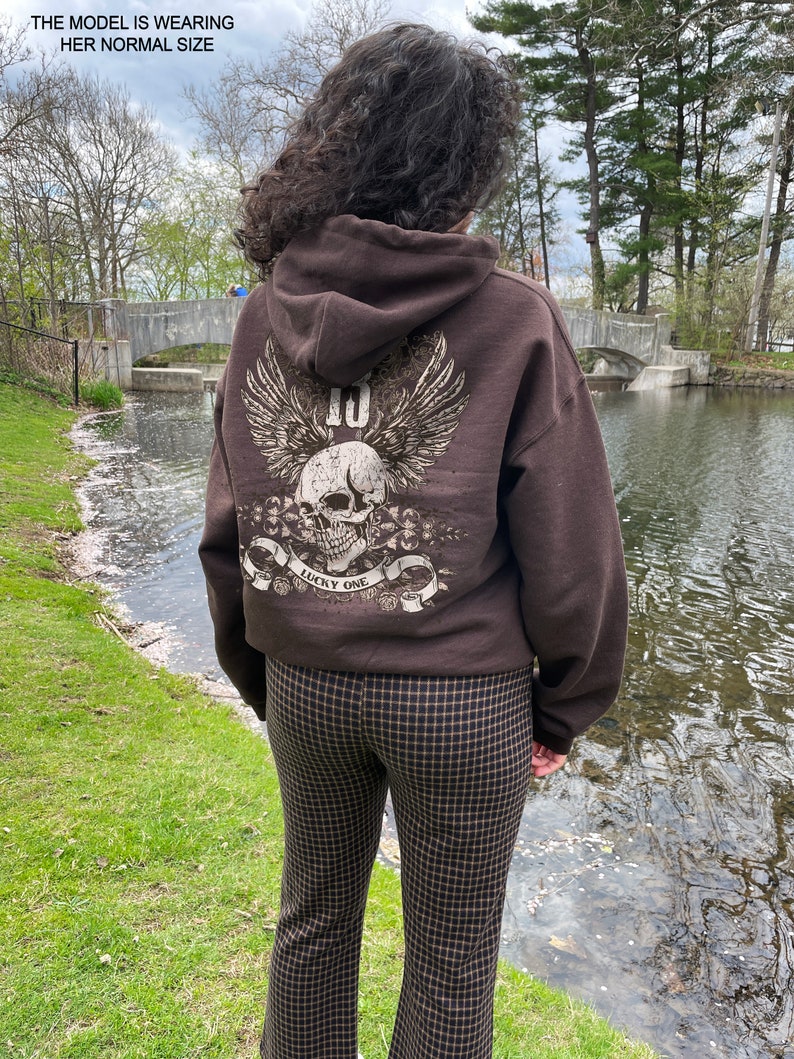 May include: Brown hooded sweatshirt with a skull graphic and the text "Lucky One" on the back. The model is wearing brown and black checkered pants.