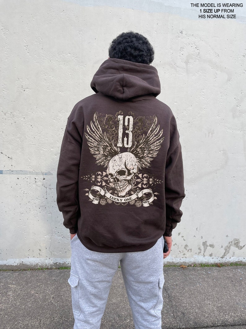 May include: Brown hooded sweatshirt with a graphic print of a skull with wings and the number 13. The text "Lucky One" is printed below the skull. The model is wearing the sweatshirt one size up from their normal size.