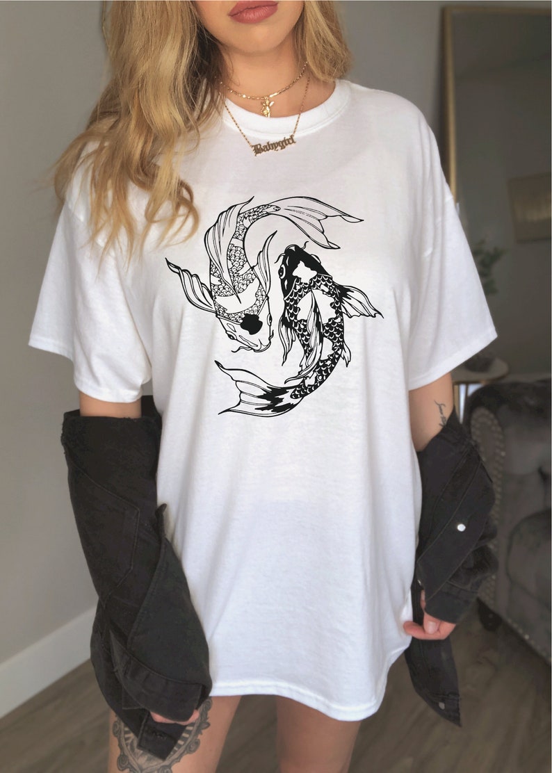 Puede incluir: Camiseta blanca de gran tama&ntilde;o con un gr&aacute;fico en blanco y negro de dos peces koi nadando en c&iacute;rculo. El texto "Babygirl" est&aacute; en un collar de cadena dorada.