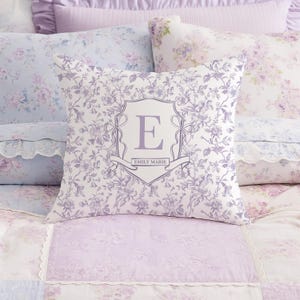 May include: A square cushion with a floral design in shades of purple and white. The cushion has a monogrammed crest with the letter "E" and the name "EMILY MARIE" on a banner. The cushion is on a bed with matching bedding.