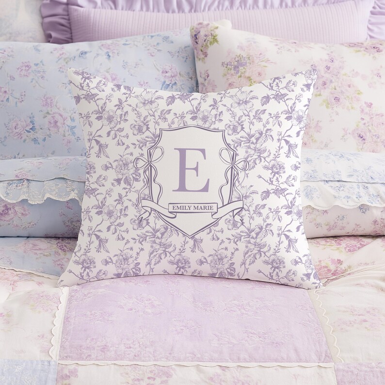 May include: A square cushion with a floral design in shades of purple and white. The cushion has a monogrammed crest with the letter "E" and the name "EMILY MARIE" on a banner. The cushion is on a bed with matching bedding.