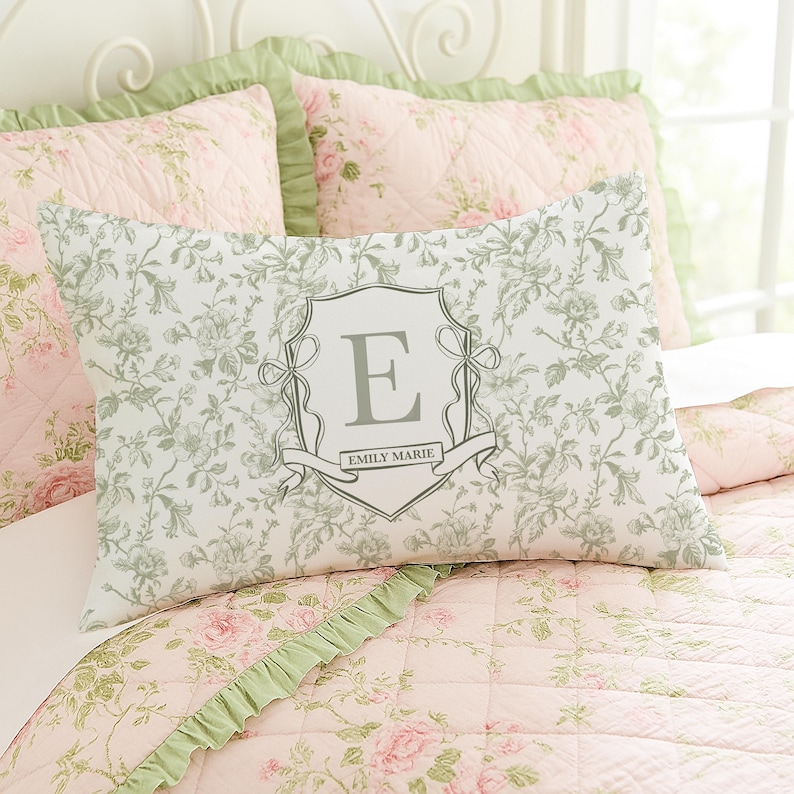 May include: A rectangular pillow with a floral pattern in shades of white and green, featuring a gray crest with the letter "E" and the name "Emily Marie". The pillow is on a bed with pink floral bedding.