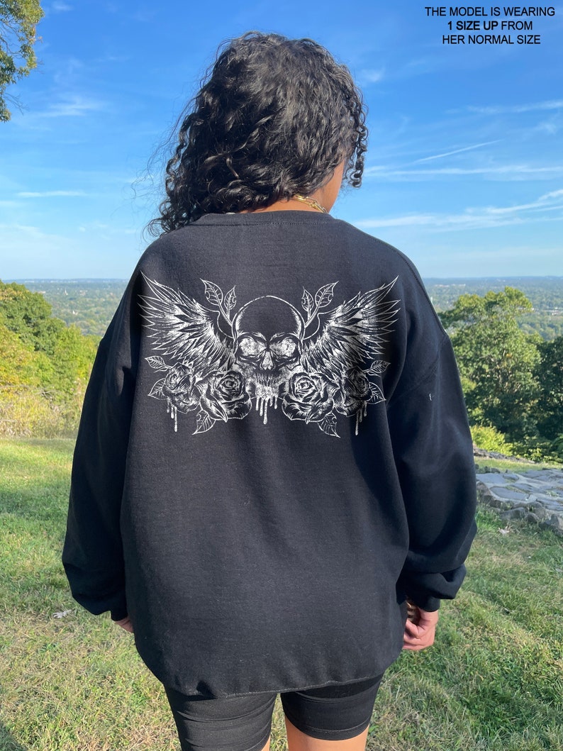 May include: Black sweatshirt with a white graphic print of a skull with wings and roses. The model is wearing the sweatshirt one size up from her normal size.