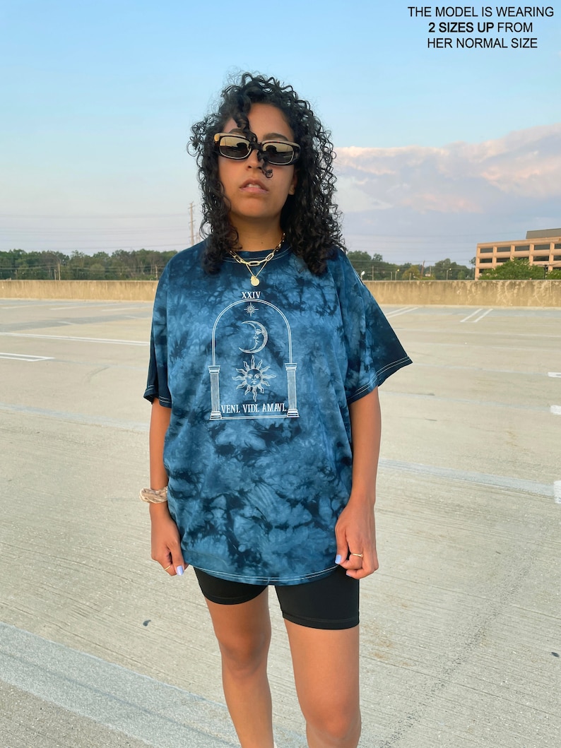 May include: A blue tie-dye t-shirt with a white graphic of an archway with a sun and moon inside. The text "XXIV" and "VENI VIDI AMAVI" are printed on the graphic. The model is wearing the shirt two sizes up from their normal size.