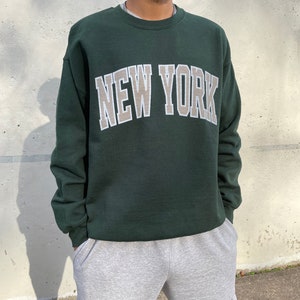 May include: A dark green sweatshirt with the text "NEW YORK" in a beige and white outline. The sweatshirt has a crew neck and long sleeves.