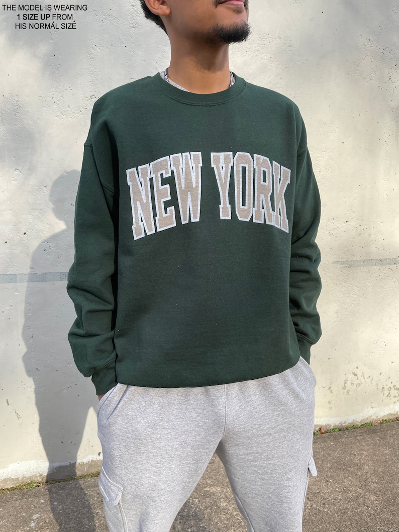 May include: A dark green sweatshirt with the text "NEW YORK" in a beige and white outline. The sweatshirt has a crew neck and long sleeves.