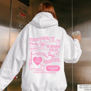 May include: White hoodie with pink text and bows. The text reads "Everything will be okay, you are his masterpiece, loved unconditionally, you are radiant in his love, you are enough in his eyes, embrace his grace within, you are loved, forgiven, redeemed, treasured soul, redeemed, you are holy in his hands, you are cherished by Christ, fearfully and wonderfully made".